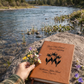 Brown leather notebook with nature-themed design on a rock by a river with mountains in the background